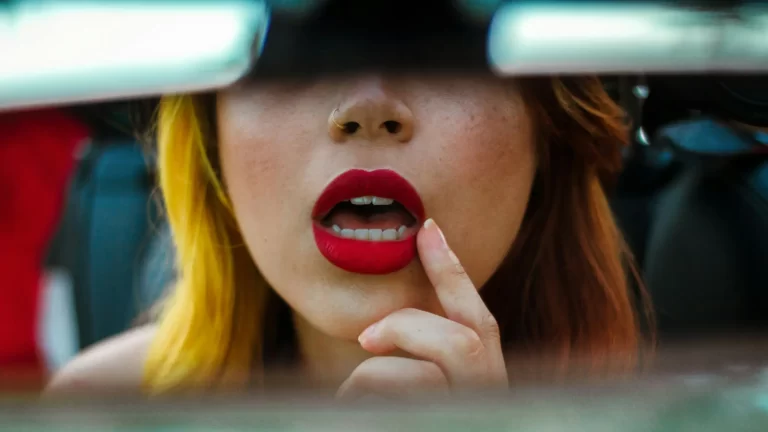 mujer viendo sus labios grandes y carnosos pintados de rojo en el espejo de su auto meses despues de aplicarse ácido hialurónico en labios