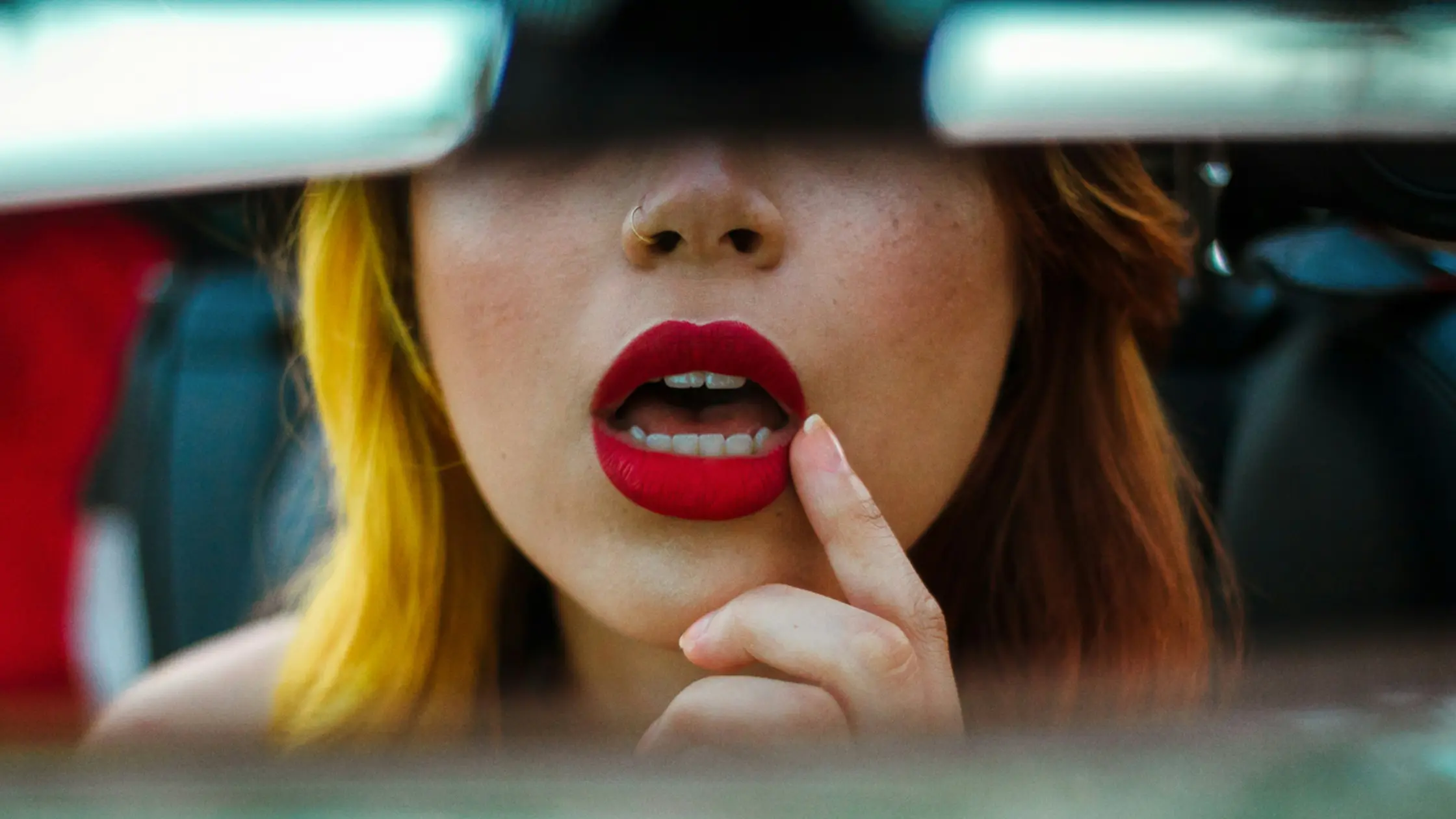 mujer viendo sus labios grandes y carnosos pintados de rojo en el espejo de su auto meses despues de aplicarse ácido hialurónico en labios